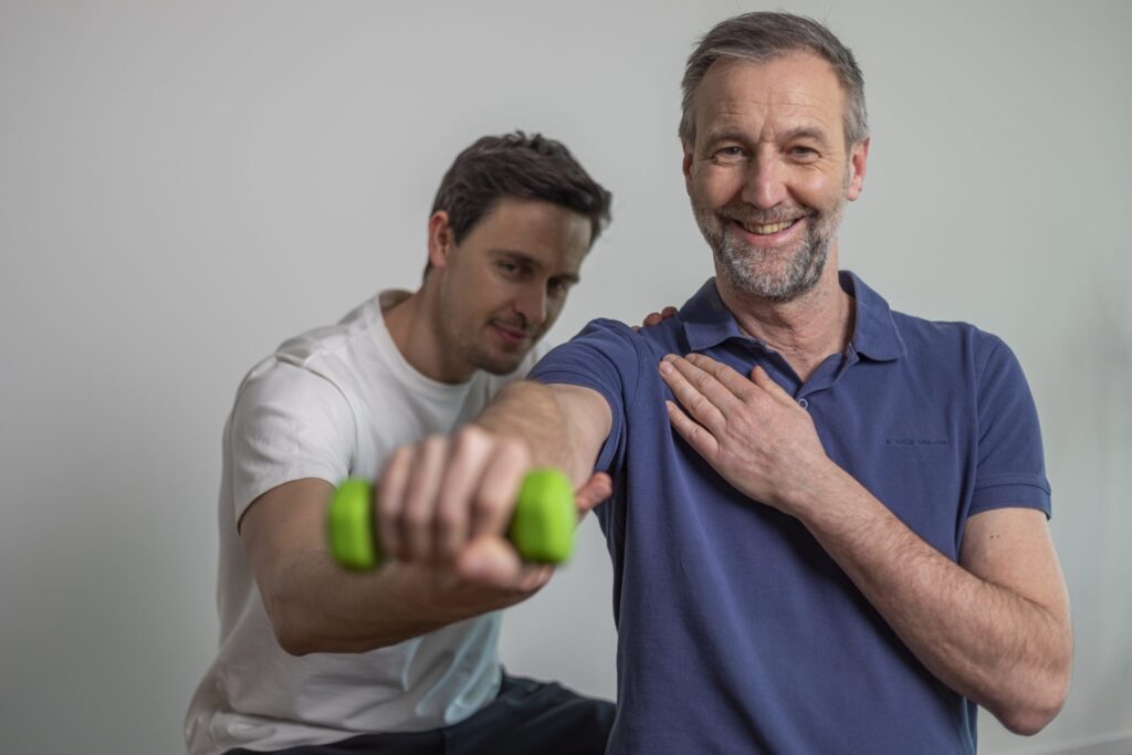 Physiotherapeut begleitet einen lächelnden Mann beim Schultertraining mit einer grünen Hantel